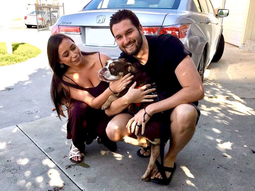A couple with a dog, smiling and posing on the driveway next to a car, holding the dog. Ends with a period.