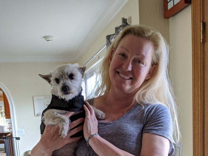 A smiling woman holds a small gray dog in her arms inside a cozy home.