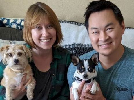A smiling couple sits on a couch holding two small dogs, one tan and fluffy, the other black and white, posing for a cozy family photo.