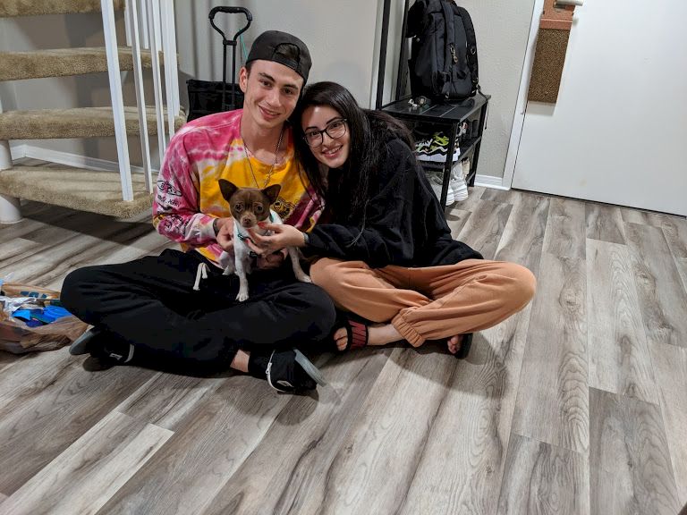 Two friends sit on a wooden floor, smiling at the camera with a small brown dog between them, in a cozy indoor space.