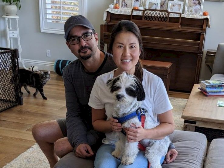 A couple sits on a couch with a small dog; a piano and cozy living room are in the background, a cat-walker gate and books nearby, smiling.