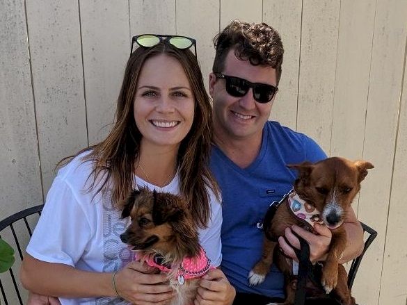 A smiling couple with two small dogs, posing outdoors against a wooden fence, both wearing sunglasses and casual clothes, cute and cozy.