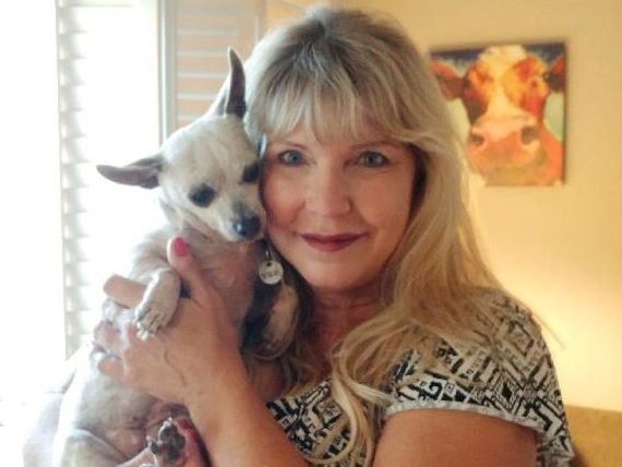 A woman with long blonde hair holds a small white and black dog close, both looking at the camera, in a cozy room.