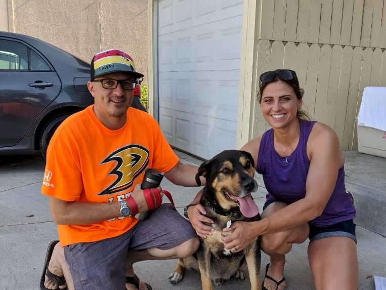 Two people with a dog outdoors, posing by a garage; man in orange shirt and cap, woman in purple top, all smiling and happy.