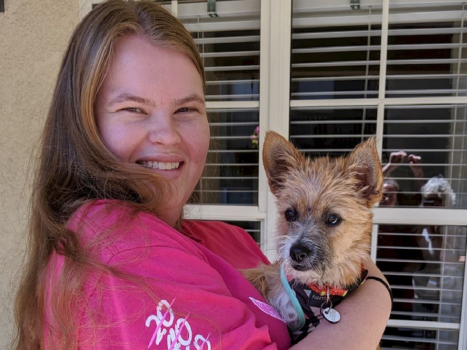 A smiling woman in a pink shirt holds a fluffy-eyed terrier mix outdoors, standing near a window with blinds, the scene warm and cheerful.