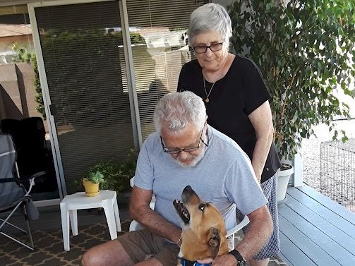 An older couple with a man sitting and a dog, outdoors on a porch; the woman stands behind him gently smiling.