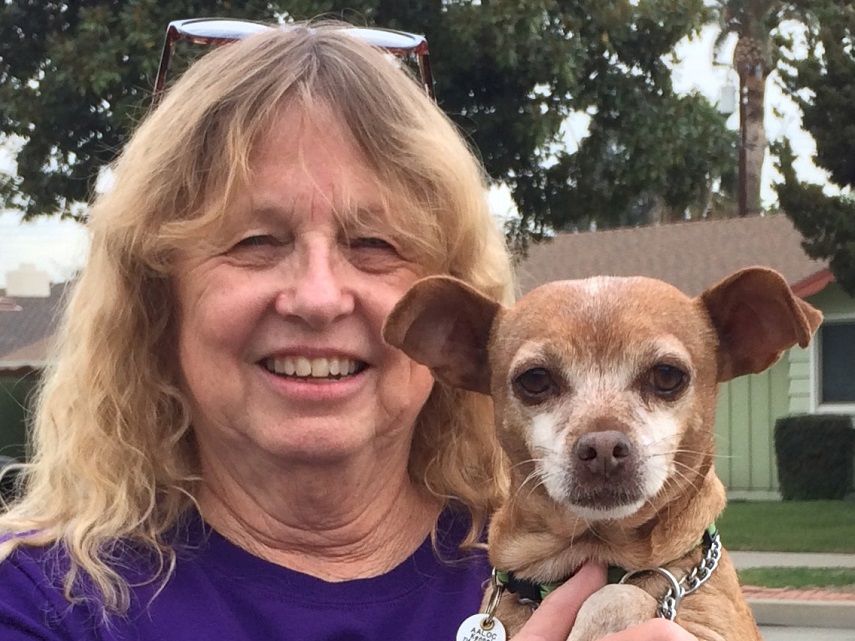 An older woman with blonde hair smiles while holding a small brown and white dog; they&rsquo;re outdoors near a house.