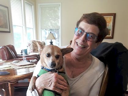 An older woman with short hair and glasses smiles while holding a light brown dog in a green sweater, sitting in a cozy living room.