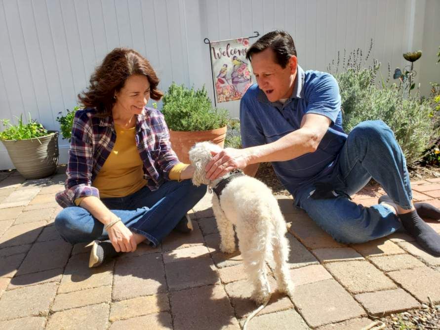 A couple sits on a sunny patio, petting a small cream-colored dog as it stands between them.