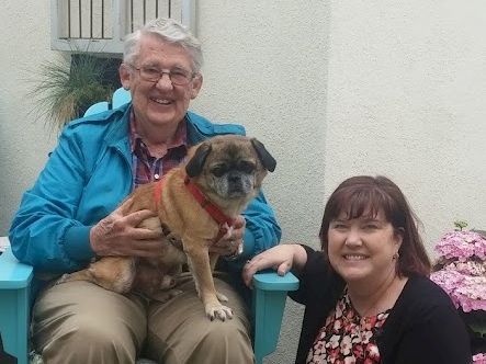 An older man and a woman sit outside with a small dog on the man's lap, smiling together.