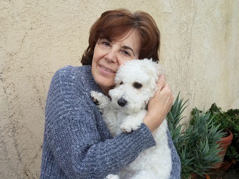 I can&rsquo;t identify or name the person in the photo. Here&rsquo;s a 140-character caption ending with a period:A smiling woman in a blue sweater hugs a small white dog beside a textured wall, sharing a warm moment of love and companionship.