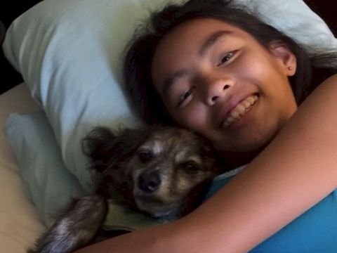A smiling person cuddles a small dog on a bed, both snuggling together under light blankets.