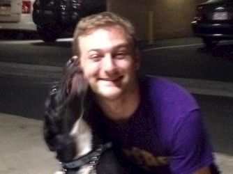 A smiling man in a purple shirt pets a black-and-white dog at night by parked cars.