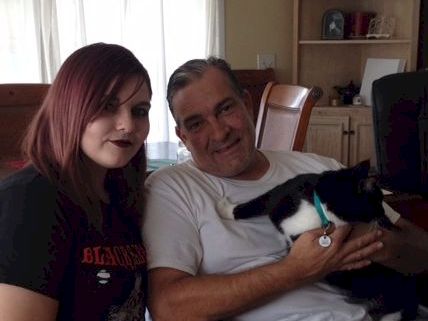 A woman and a man sit together indoors, smiling, with a small black-and-white cat on the man&rsquo;s lap.