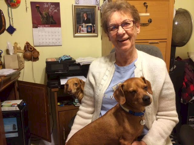 An elderly woman smiles while sitting with two brown dogs in a cozy, cluttered home office.