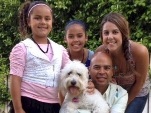 A family portrait with two girls, a woman, a man, and a fluffy white dog posing outdoors in front of a fence and greenery.