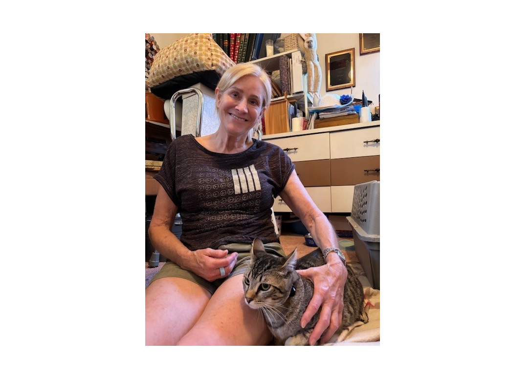 A smiling woman sits on the floor with a tabby cat on her lap, in a cozy, cluttered room with shelves and furniture around.